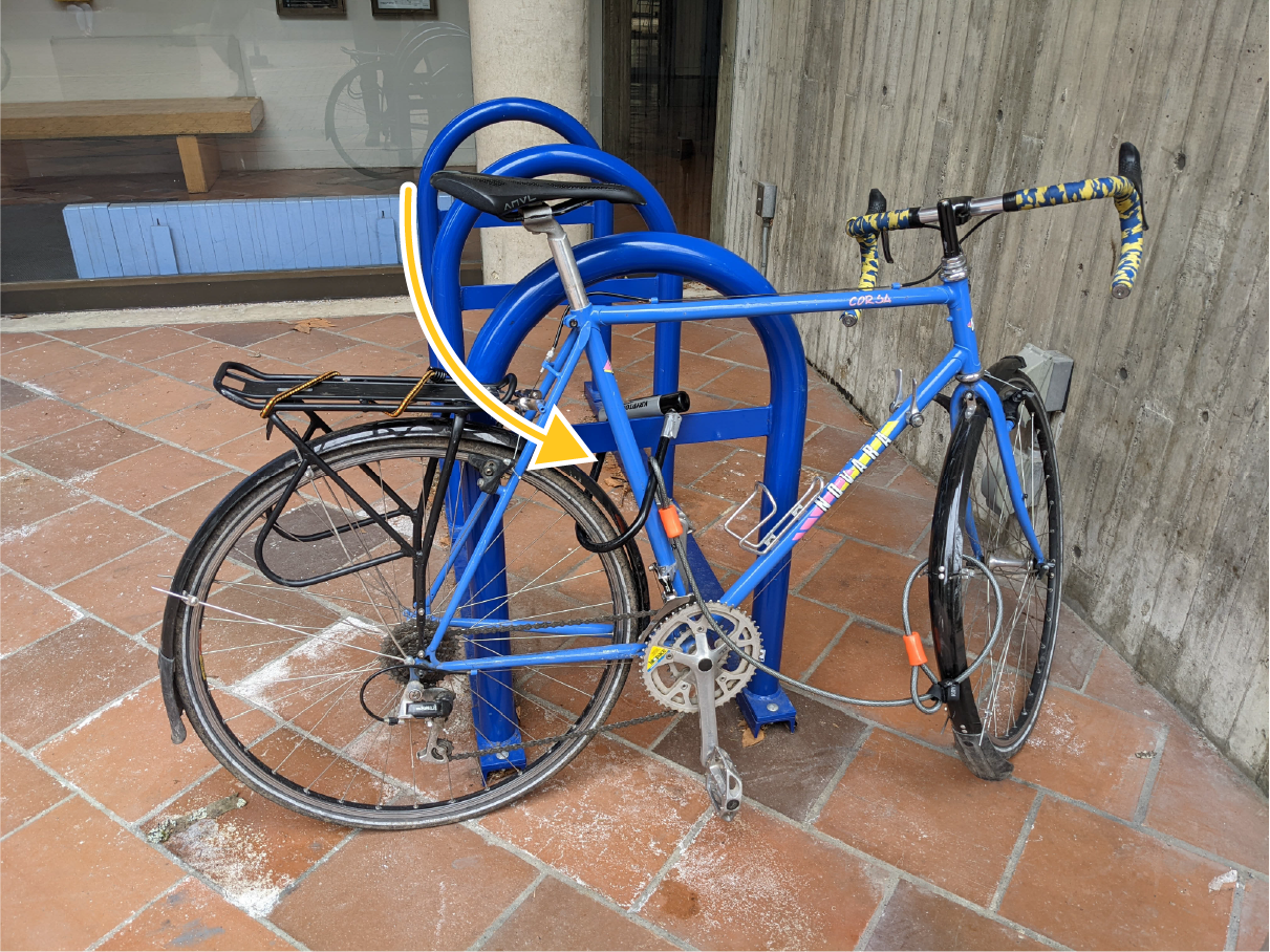 A blue bike locked to a rack using a u-lock around the frame and the rear wheel. A cable connects the front wheel to the u-lock.