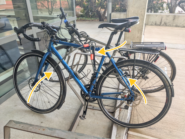 A modern blue bike locked to a rack using a u-lock on the frame and the rack and a cable around both the front and rear wheels connected to the u-lock.